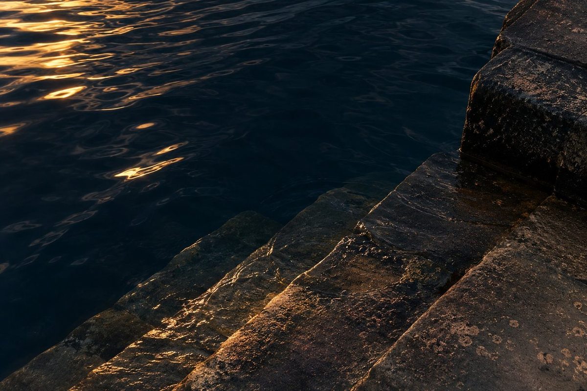 Old stone harbour steps descending into calm water at golden hour, with the waterline marked on the stone